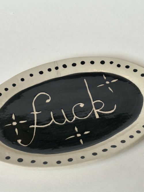 Assiette ovale noire avec texte 'fuck' doré, liseré de pois et détails étoiles.