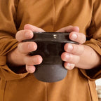 Mug noir brillant tenu par les deux mains, vue du bord et de l’anse
