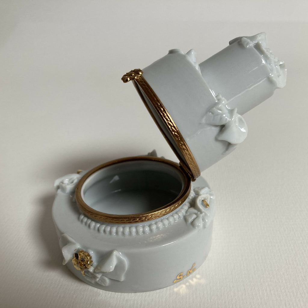 Boîte en porcelaine blanche ouverte, vue de biais, intérieur sombre et filet doré.