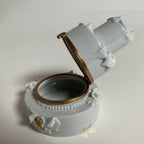 Boîte en porcelaine blanche ouverte, vue de biais, intérieur sombre et filet doré.