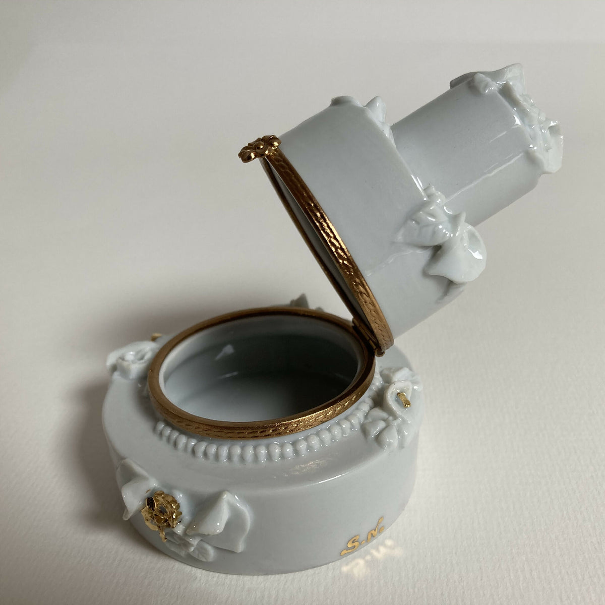 Boîte en porcelaine blanche ouverte, vue de biais, intérieur sombre et filet doré.