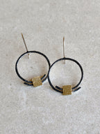 Boucles d’oreilles pendantes : deux anneaux noirs avec carrés dorés texturés, tiges argentées.