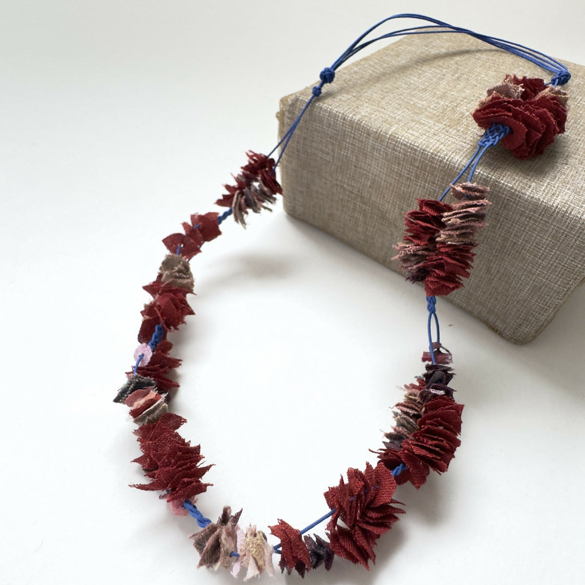 Collier en cordon bleu avec fleurs en tissu bordeaux, vue de côté sur boîte