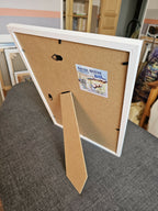Dos de tableau encadré en carton, étiquette et attache murale visibles.