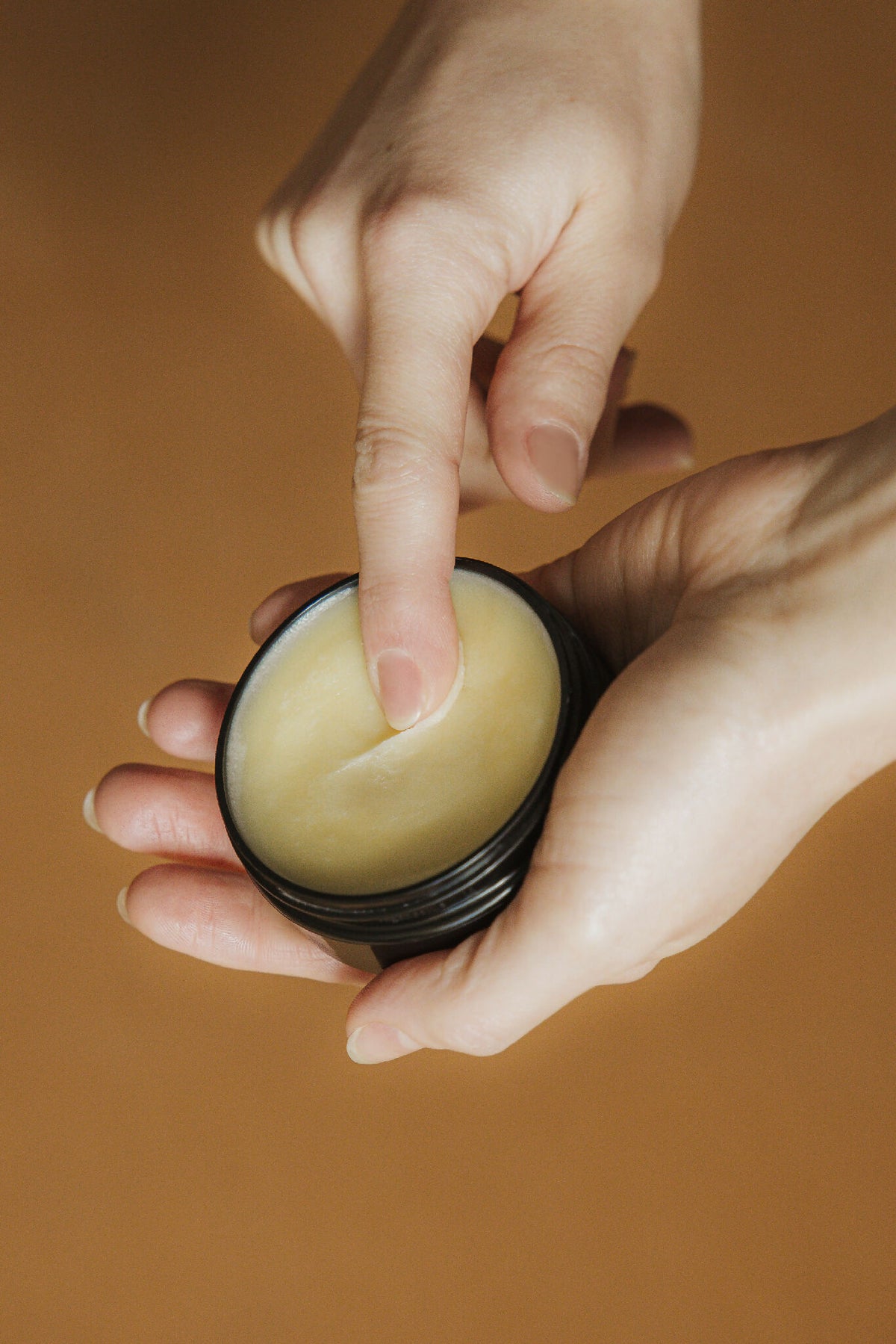 Baume noisette dans un petit pot noir, mains tenant le produit, texture jaune pâle.
