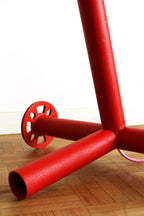 Pied de lampe rouge avec tubes horizontaux et disque de fixation, sol en bois