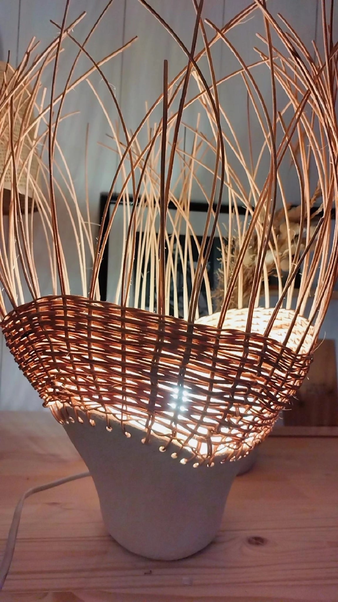Luminaire en rotin brun tressé sur pot en grès, éclairage chaud diffus.