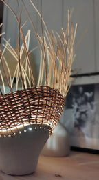 Luminaire en rotin brun tressé sur pot en grès, bord ajouré et lumière chaude.