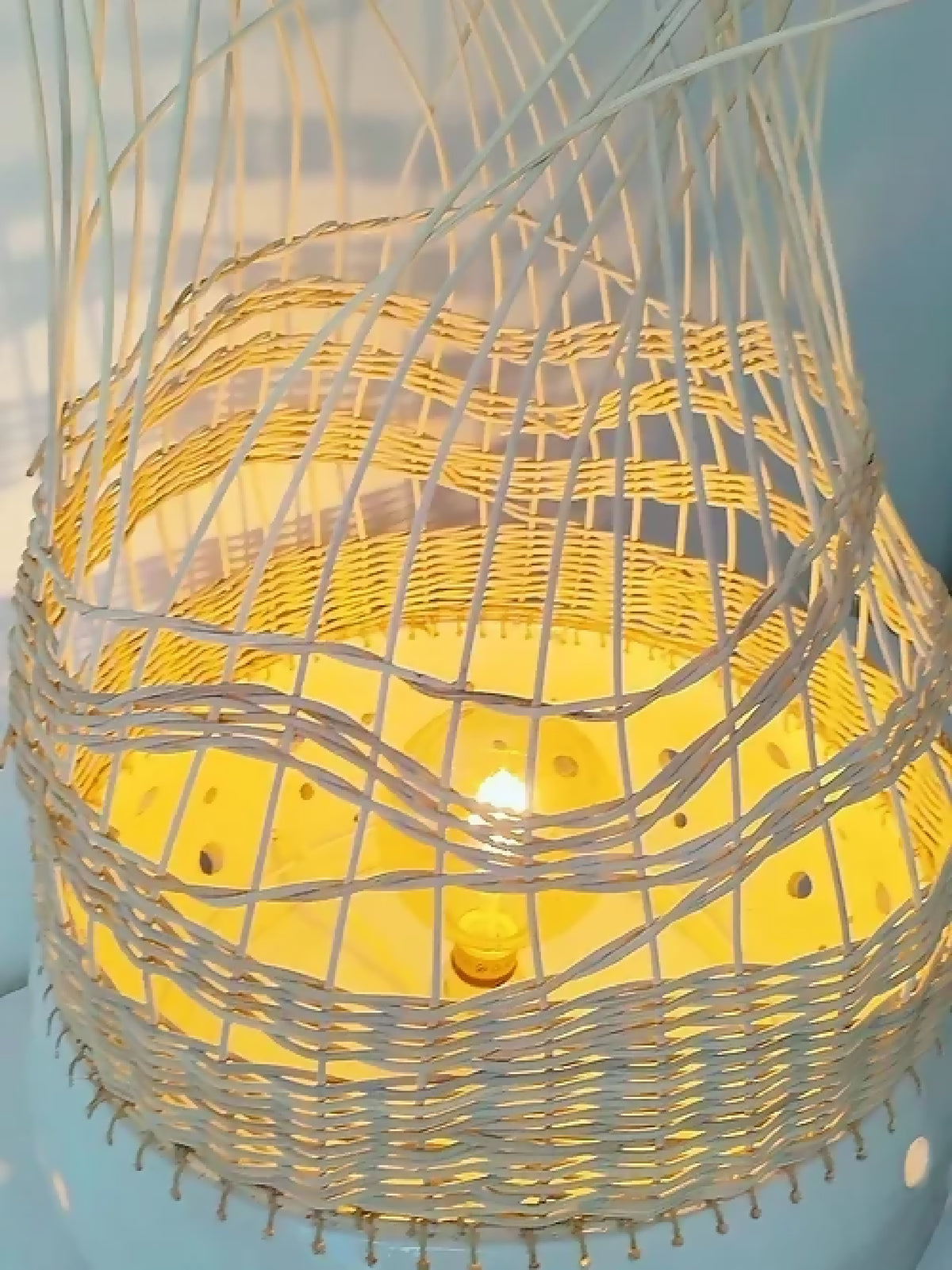 Lampe à poser tressée blanc et jaune, abat-jour grillagé, lumière allumée.