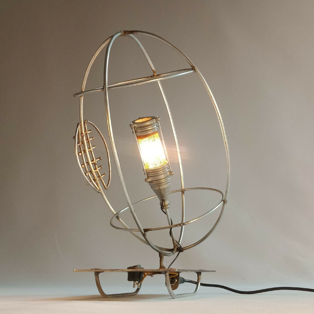 Lampe RUGBY allumée : cage ronde dorée, ampoule éclairante et base sur pied