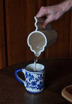 Vase blanc tenu à la main, versement d’un liquide dans une petite tasse bleue.