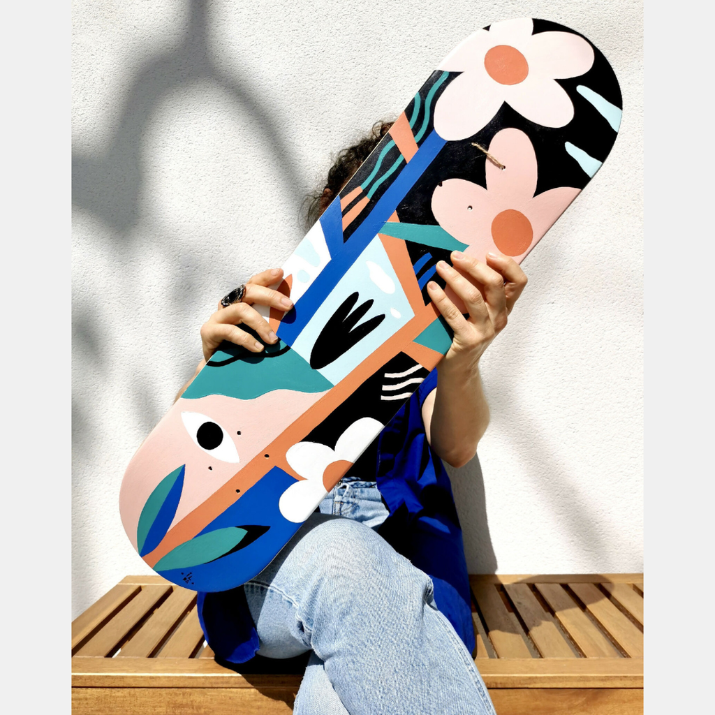 Skateboard illustré, motifs de visages et fleurs, couleurs vives bleu, orange et noir.