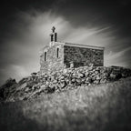Photographie noir et blanc d’une chapelle en pierre sur une colline, ciel dramatique.