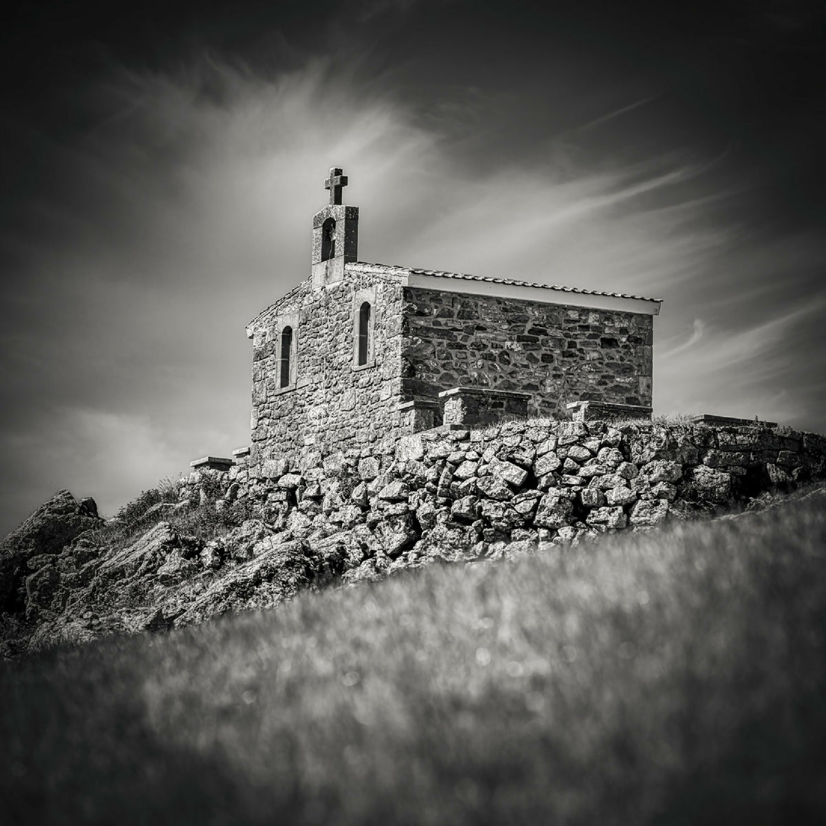 Photographie noir et blanc d’une chapelle en pierre sur une colline, ciel dramatique.