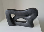 Sculpture abstraite sombre aux courbes sinueuses, trois ouvertures ovales visibles.