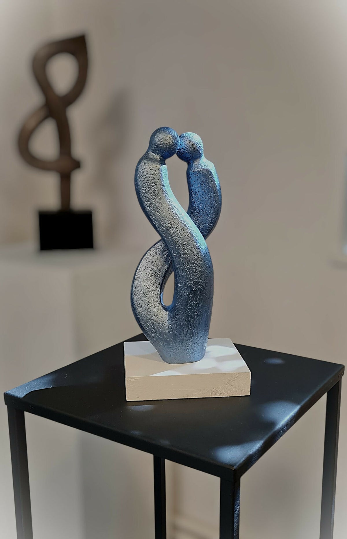 Sculpture couple abstrait en métal bleu sur socle blanc, posée sur table noire.