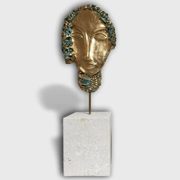 Sculpture de tête en métal doré sur socle rectangulaire blanc, tresse et visages stylisés