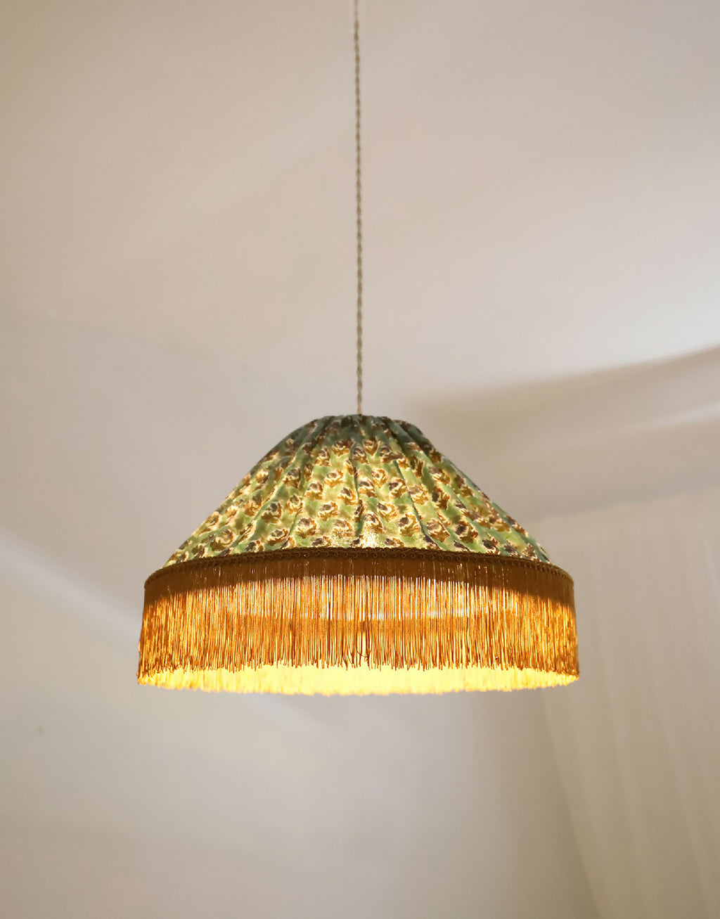 Abat-jour de suspension vu de côté : tissu doré-vert à motifs, franges dorées.