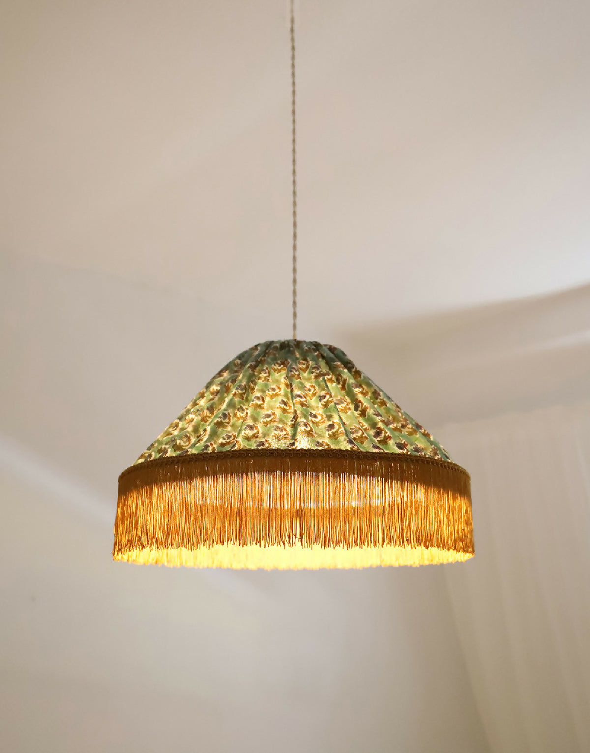 Abat-jour de suspension vu de côté : tissu doré-vert à motifs, franges dorées.