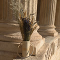 Vase en céramique beige avec anse et fleurs séchées, posé sur un socle.