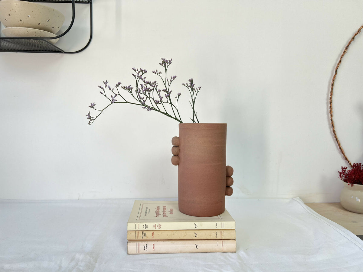 Vase en grès brun cylindrique à poignées, bouquet violet posé sur des livres.